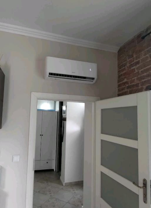 Biały klimatyzator ścienny w jasnym pokoju z ceglanym akcentem, widoczny fragment białych drzwi z przeszkleniami i wnętrze szafy w tle.