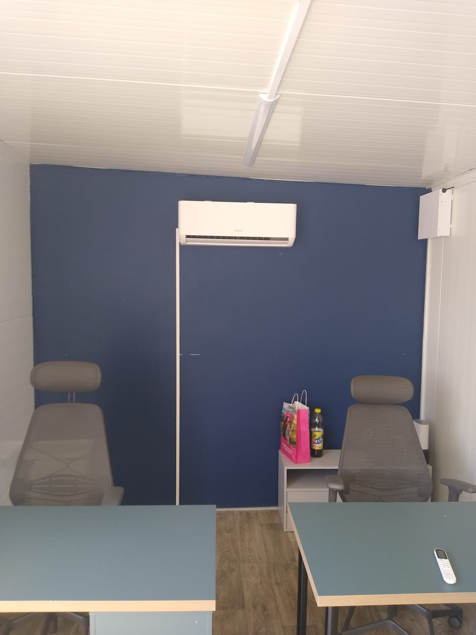 Wnętrze biurowe w kontenerze z zainstalowanym klimatyzatorem na niebieskiej ścianie, dwa krzesła biurowe, biurka, torba na zakupy i butelka na stoliku, pilot do klimatyzacji na biurku.