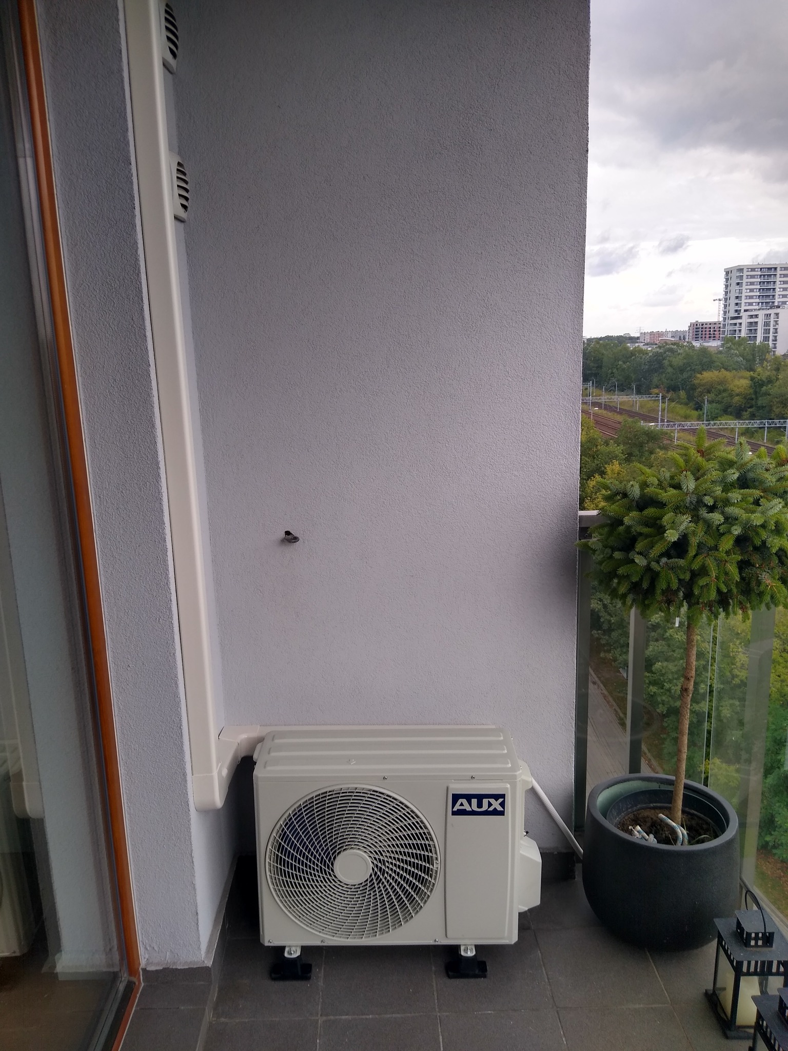 Jednostka zewnętrzna klimatyzatora AUX zamontowana na balkonie z widokiem na drzewa i budynki w tle; obok doniczka z małym drzewkiem bonsai.