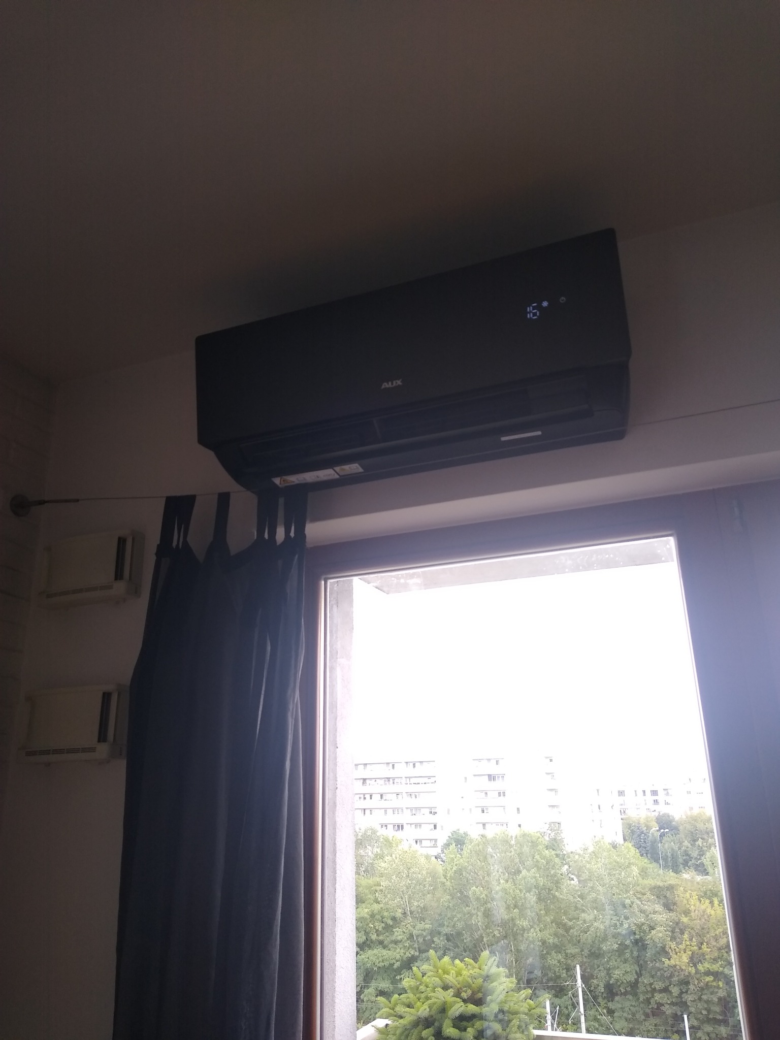 Czarny klimatyzator AUX zamontowany nad oknem z widokiem na budynki i drzewa, w tle zasłony zaciemniające.