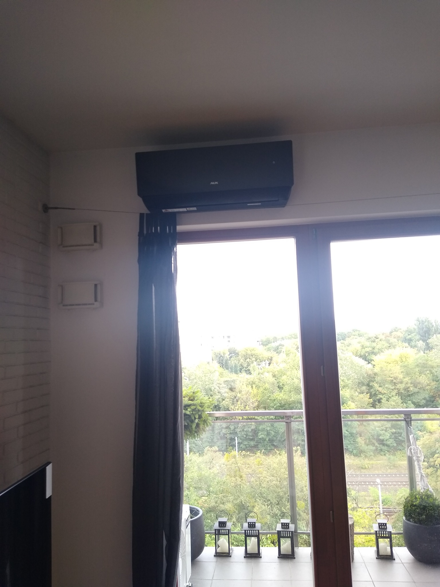 Czarny klimatyzator na ścianie w mieszkaniu z widokiem na zieleń z balkonu, na którym stoją lampiony i donica z rośliną.