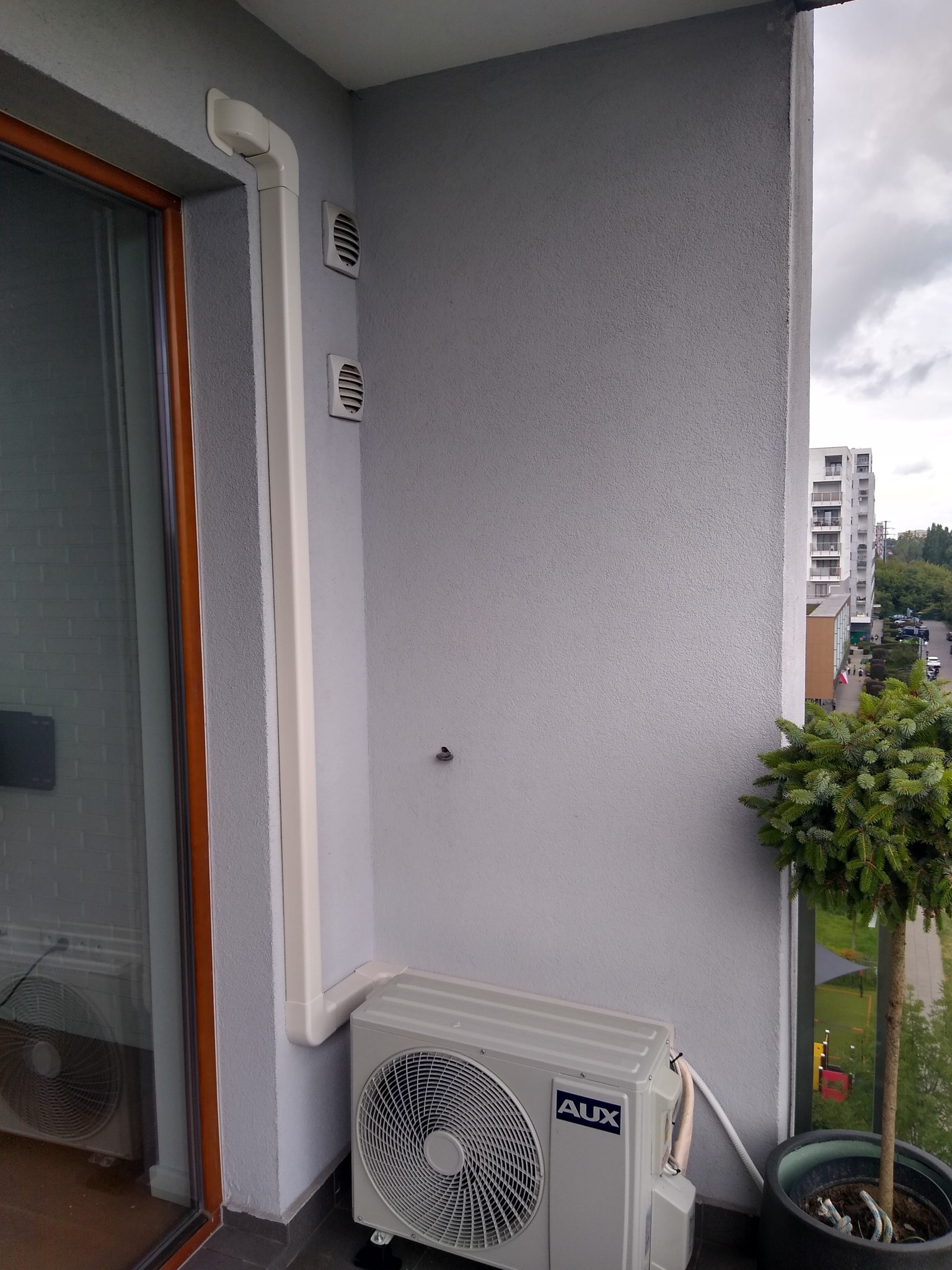 Jednostka zewnętrzna klimatyzatora marki AUX zamontowana na balkonie bloku mieszkalnego, widoczne rury odprowadzające skropliny i kratki wentylacyjne na ścianie.