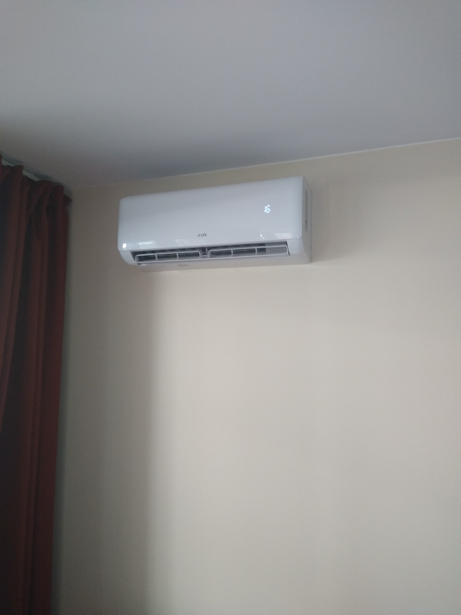 Biały klimatyzator AUX zamontowany na jasnej ścianie, wyświetlający temperaturę 16 stopni, z zasłoną w tle.