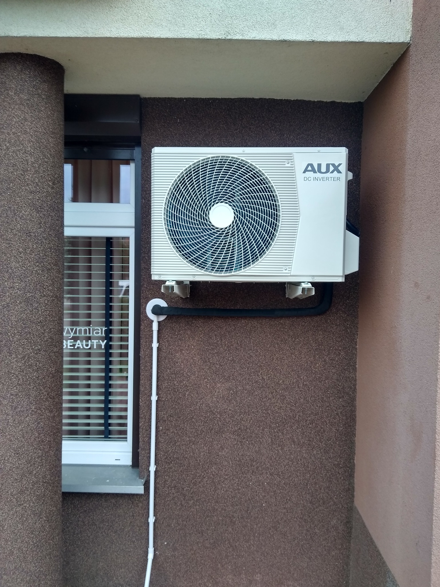 Zewnętrzna jednostka klimatyzacji AUX DC Inverter zamontowana na brązowej, chropowatej ścianie budynku obok okna z napisem 'Wymiar Beauty'.