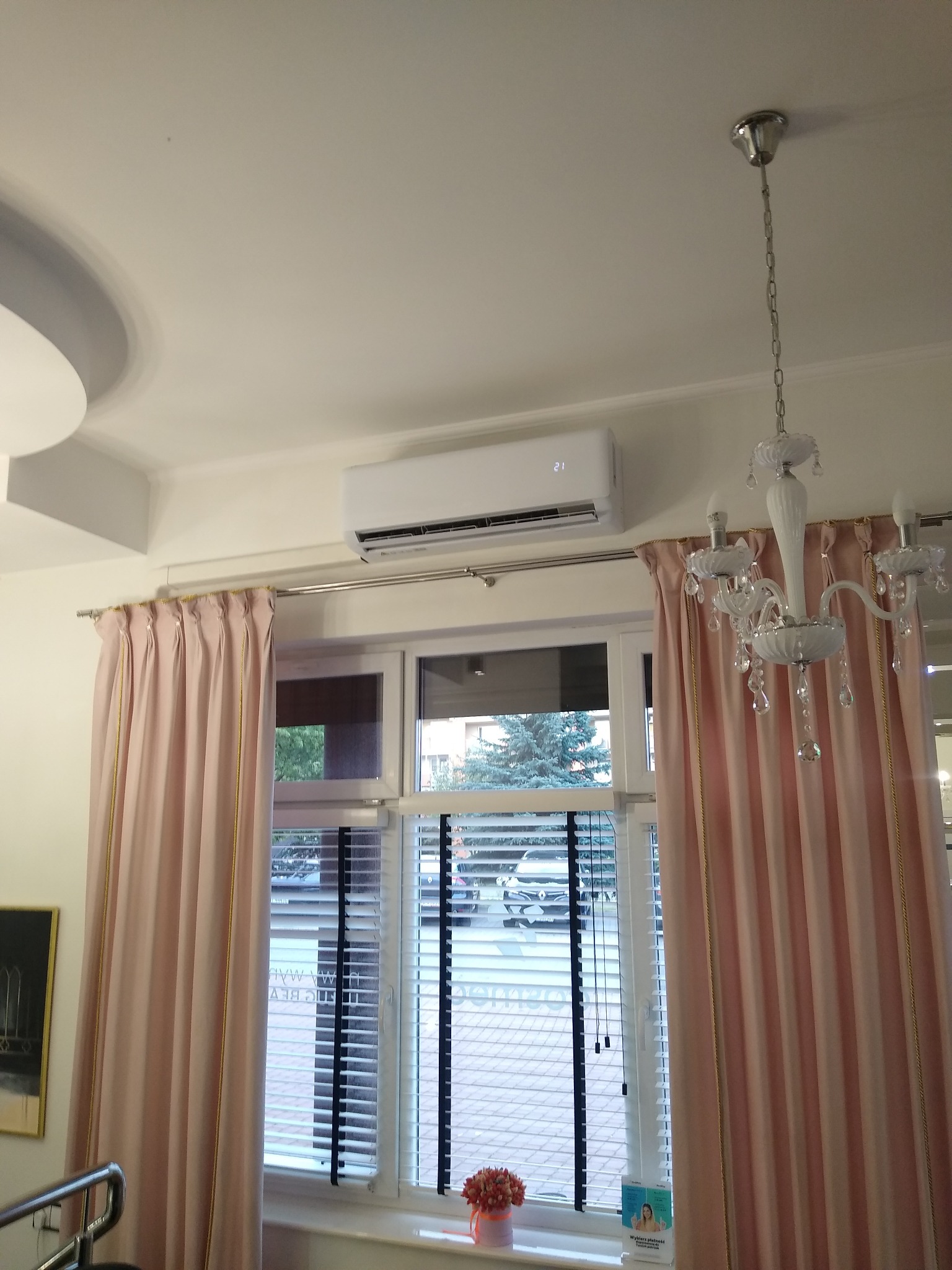 Biała jednostka klimatyzacyjna zamontowana nad oknem z różowymi zasłonami i żaluzjami, widoczny również ozdobny żyrandol.