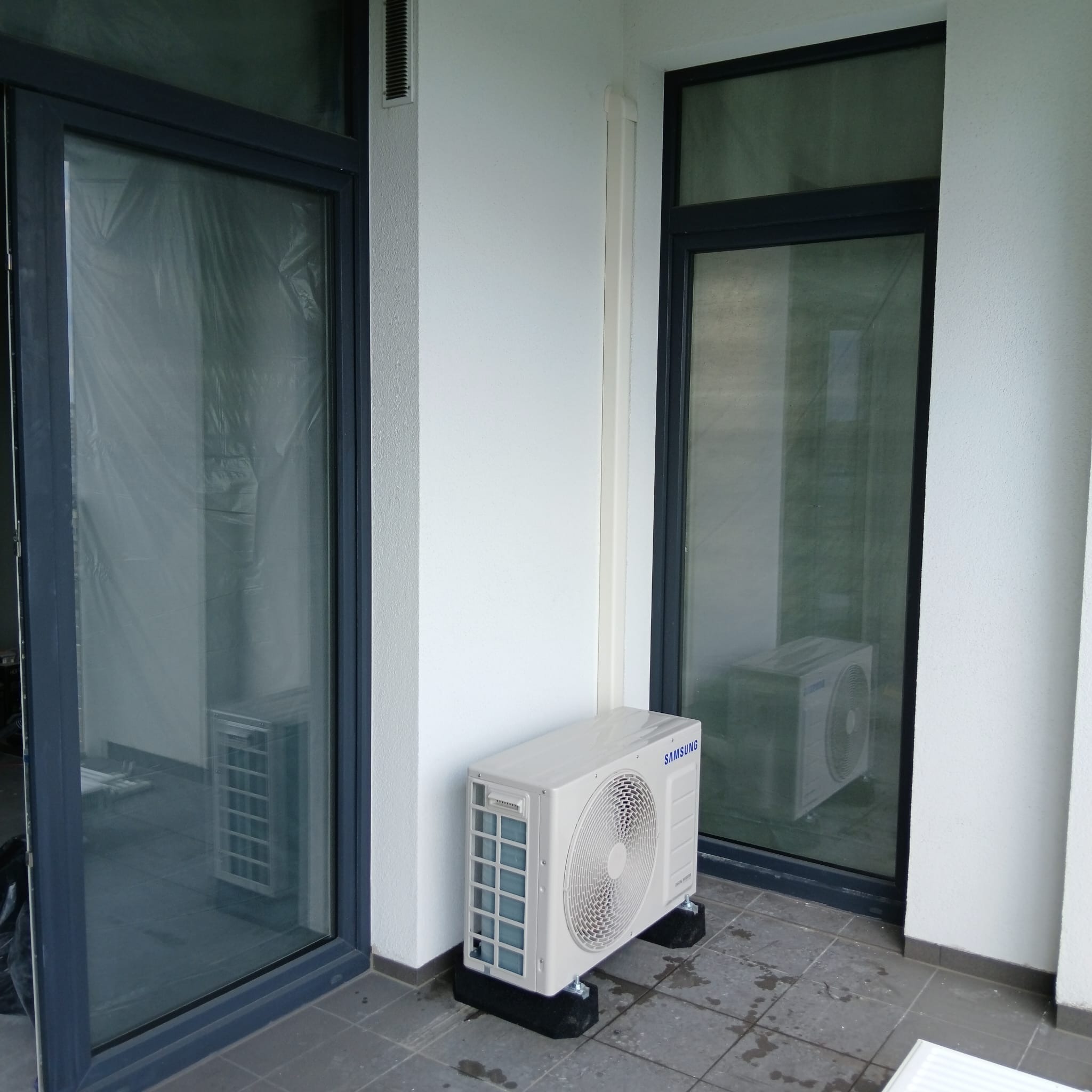 Biała jednostka zewnętrzna klimatyzatora Samsung na czarnych podkładkach antywibracyjnych, zamontowana na szarym balkonie z widokiem na okna zabezpieczone folią ochronną.