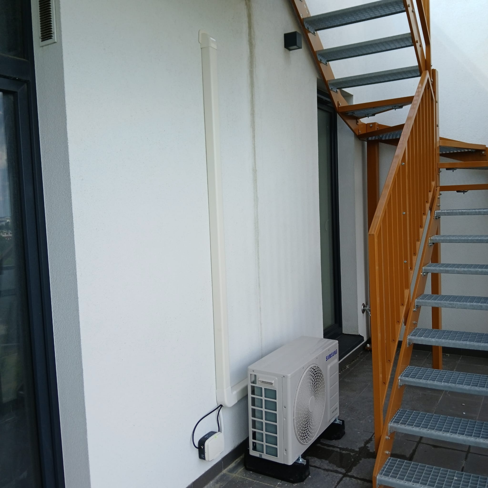 Zewnętrzna jednostka klimatyzacji Samsung zamontowana na balkonie z metalowymi schodami, widoczne przewody i listwa maskująca na ścianie.