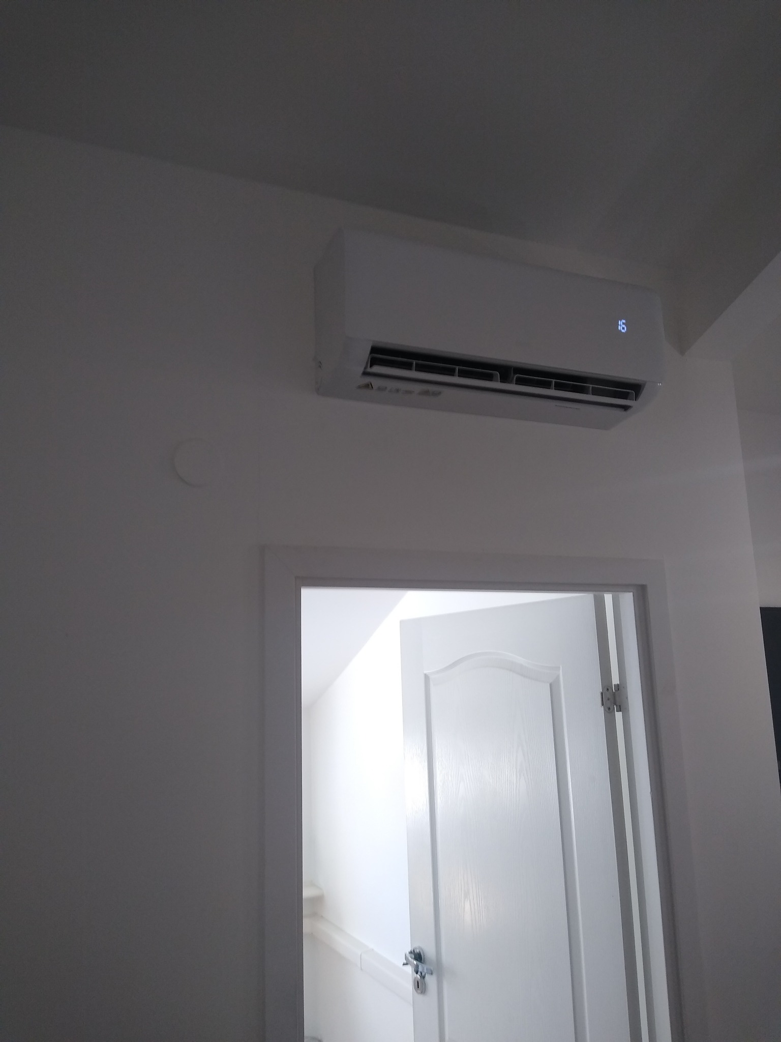 Biały klimatyzator ścienny zamontowany nad otwartymi drzwiami do jasnego pomieszczenia. Na ścianie widoczny okrągły ślad po usuniętym elemencie.