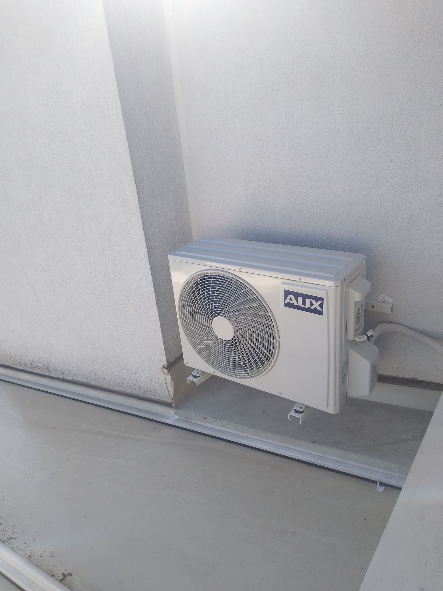 Zewnętrzna jednostka klimatyzacji marki AUX zamontowana na białej, teksturowanej ścianie budynku. Widoczne metalowe wsporniki i rury odprowadzające.
