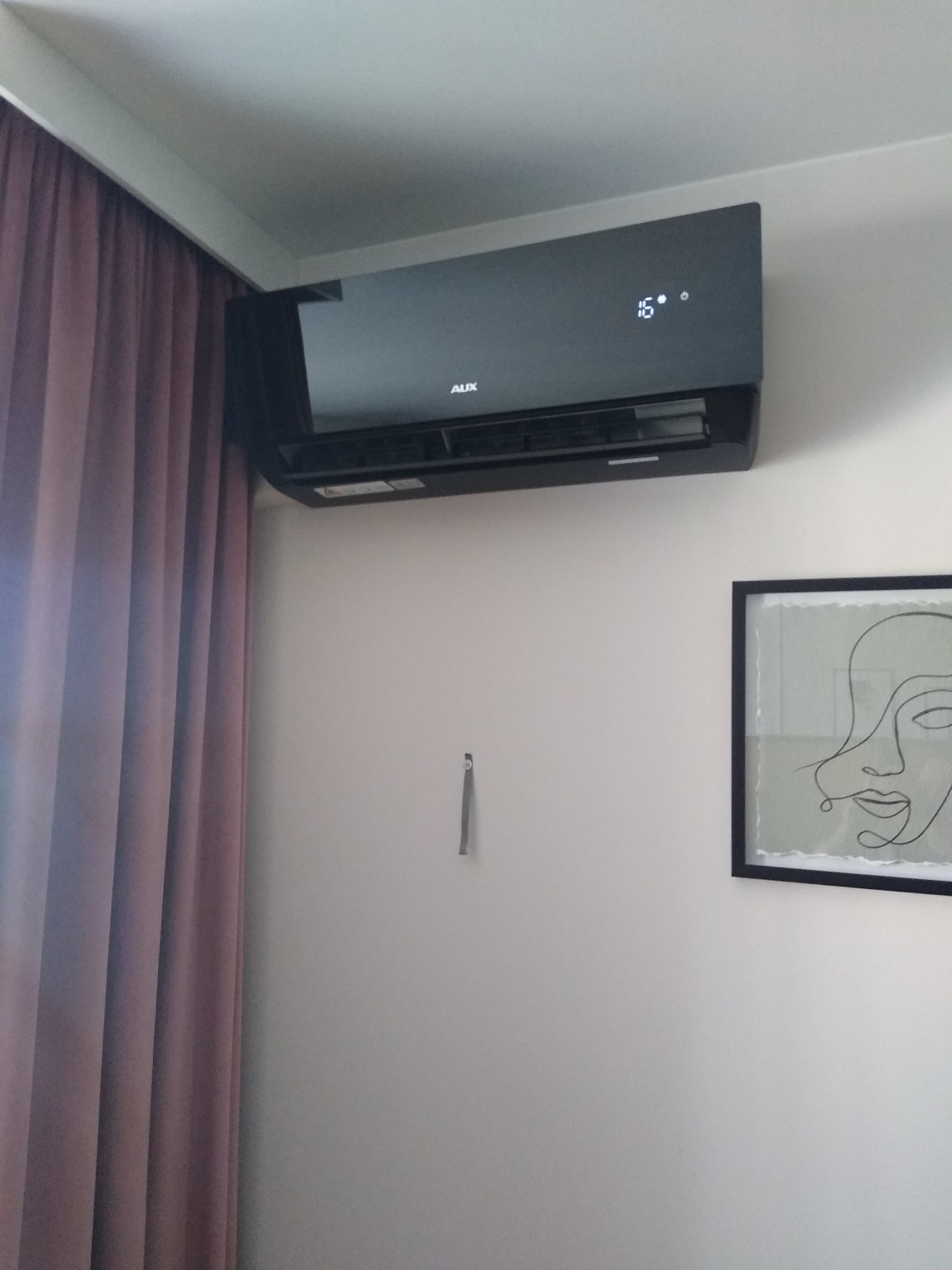 Czarny klimatyzator AUX zamontowany na białej ścianie w pokoju z zasłoną i obrazem w ramie.