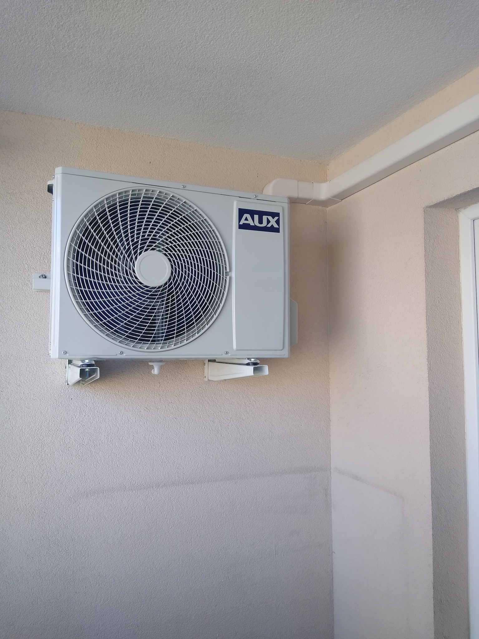 Zewnętrzna jednostka klimatyzacji AUX zamontowana na beżowej ścianie budynku, z widocznymi białymi rurami odprowadzającymi.