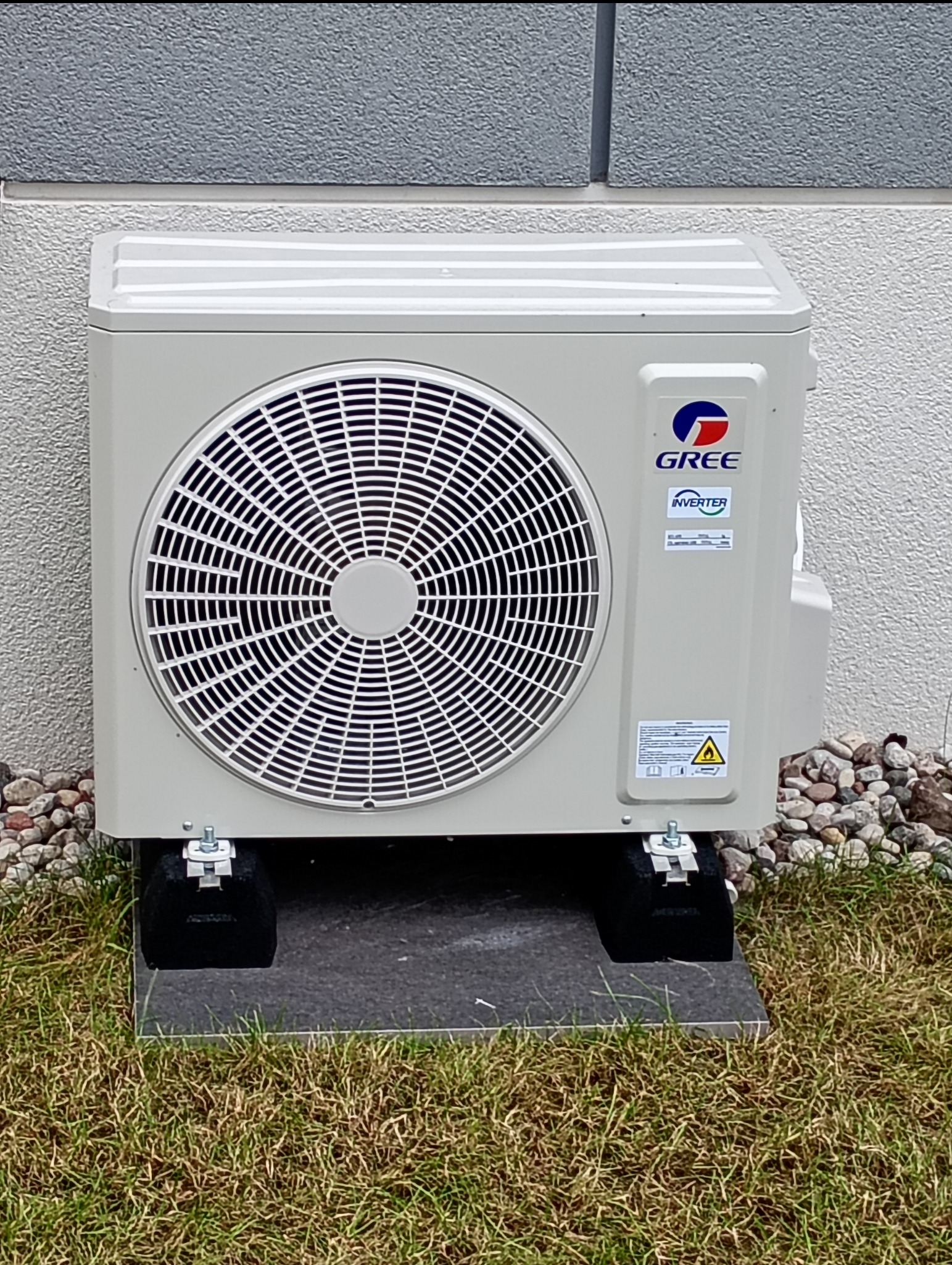 Zewnętrzna jednostka klimatyzacji marki Gree, zamontowana na betonowej płycie i gumowych podkładkach, na tle ściany budynku i trawnika.
