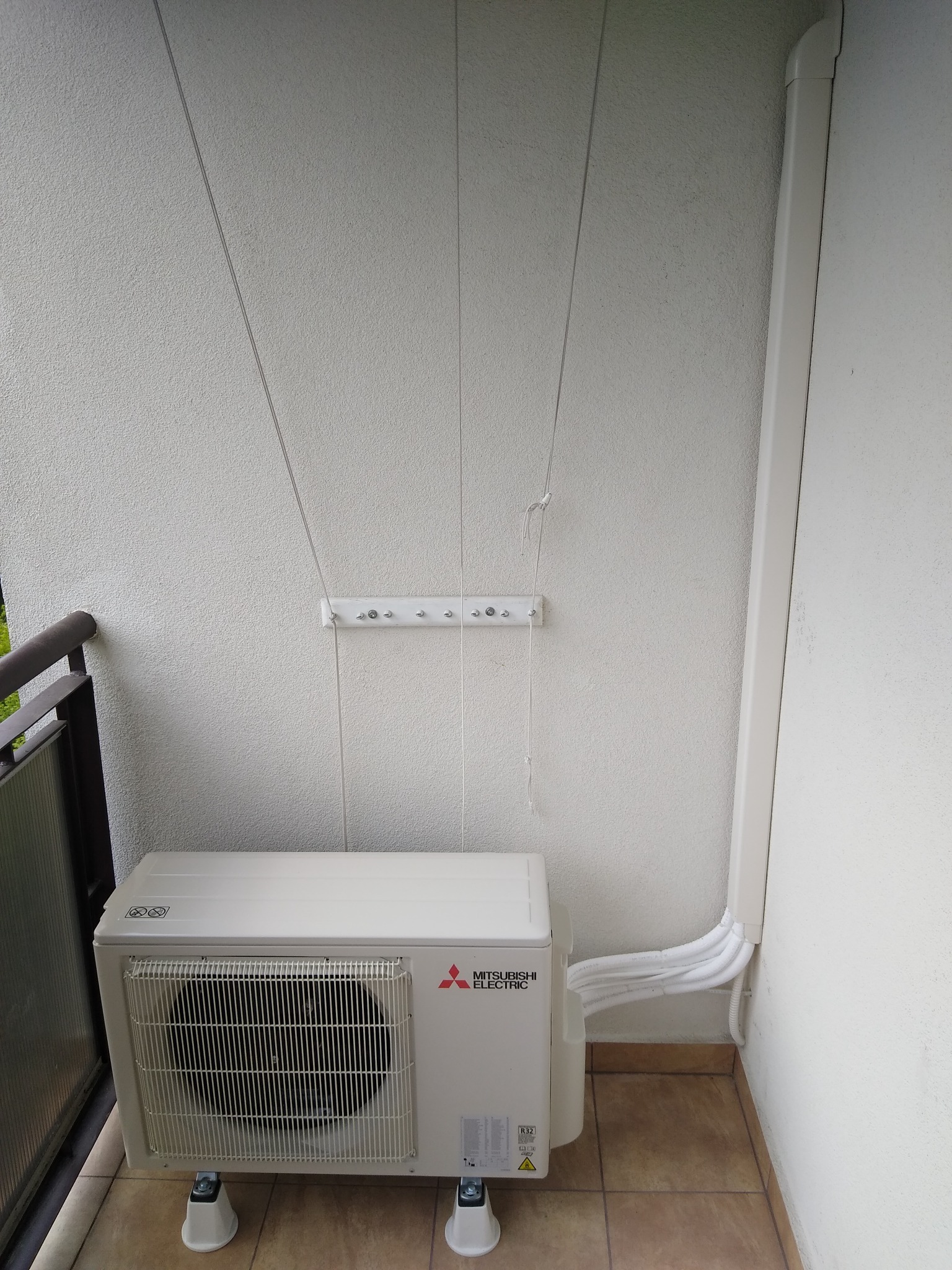 Klimatyzator Mitsubishi Electric na balkonie w Warszawie, zamontowany na podwyższeniu. Widoczne rurki odprowadzające. Ściana o jasnej strukturze.
