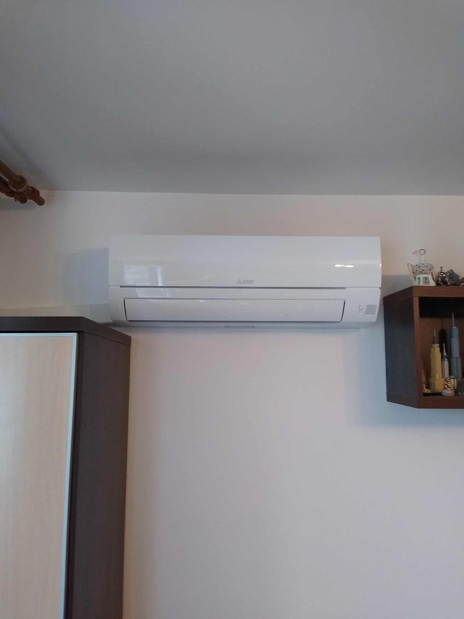 Biała klimatyzacja Mitsubishi Electric zamontowana na ścianie w pomieszczeniu, widoczny fragment szafy i półki z dekoracjami. Ujęcie z dołu.