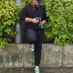 Mężczyzna w czarnym ubraniu i czapce, z profesjonalnym aparatem fotograficznym w rękach, oparty o betonowy murek obok krzewu.