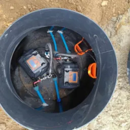 Wnętrze studni głębinowej z dwoma pompami i sterownikami, widoczne niebieskie rury i elementy instalacji, otoczone czarnym tworzywem sztucznym, z piaskiem na zewnątrz.