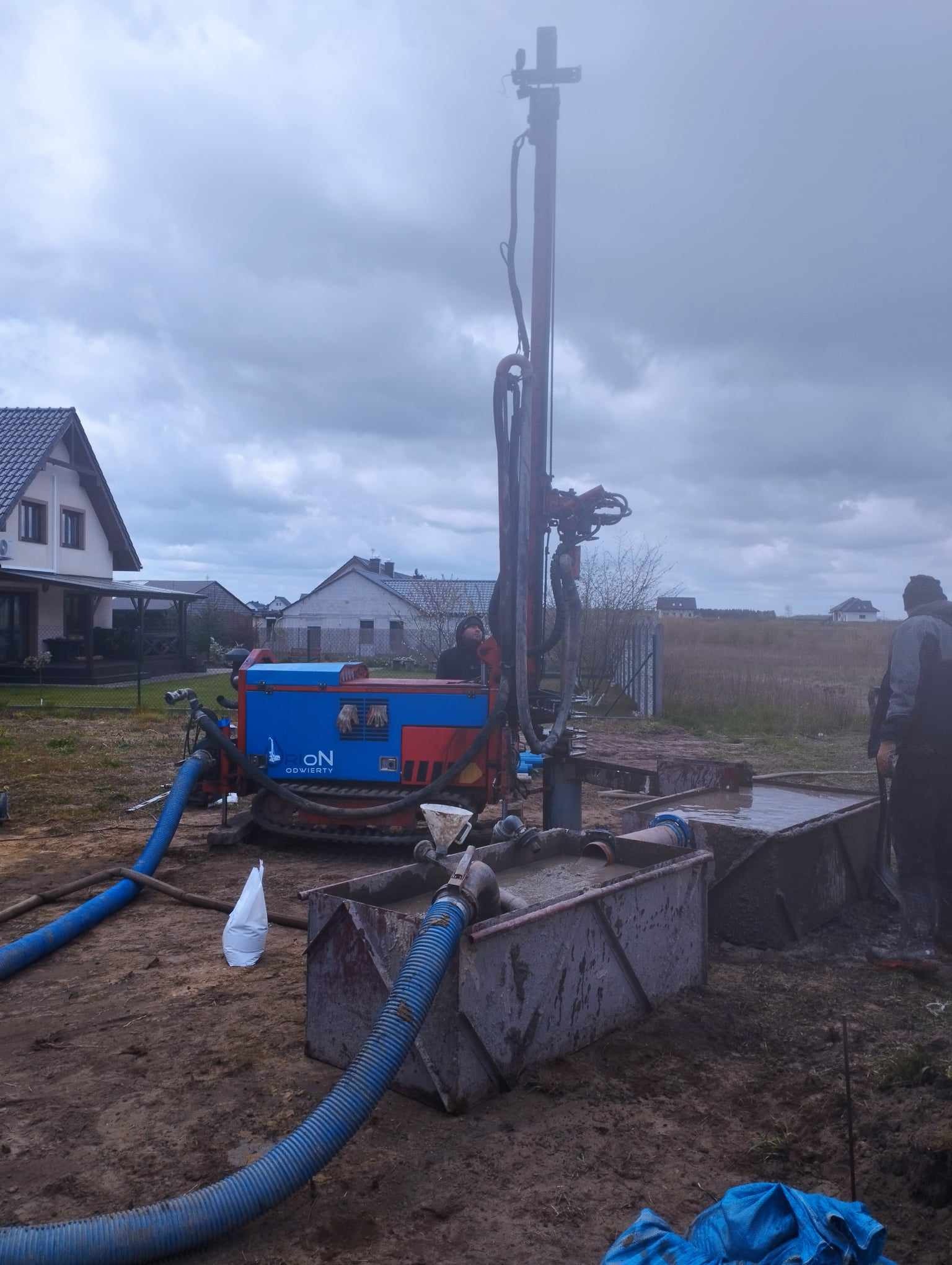 Wiertnica na gąsienicach z logo 'ON ODWIERTY' podczas pracy na błotnistym terenie, widoczne dwa zbiorniki z szarą cieczą, w tle domy jednorodzinne i pochmurne niebo.