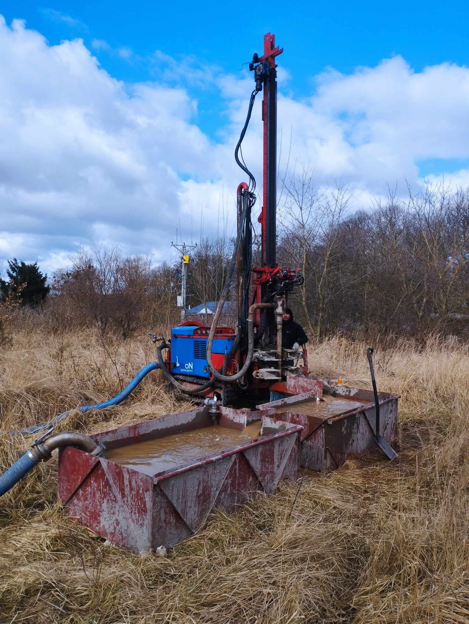 Wiertnica w trakcie pracy na zewnątrz, woda w zbiornikach, operator w kabinie, wysoka trawa dookoła.