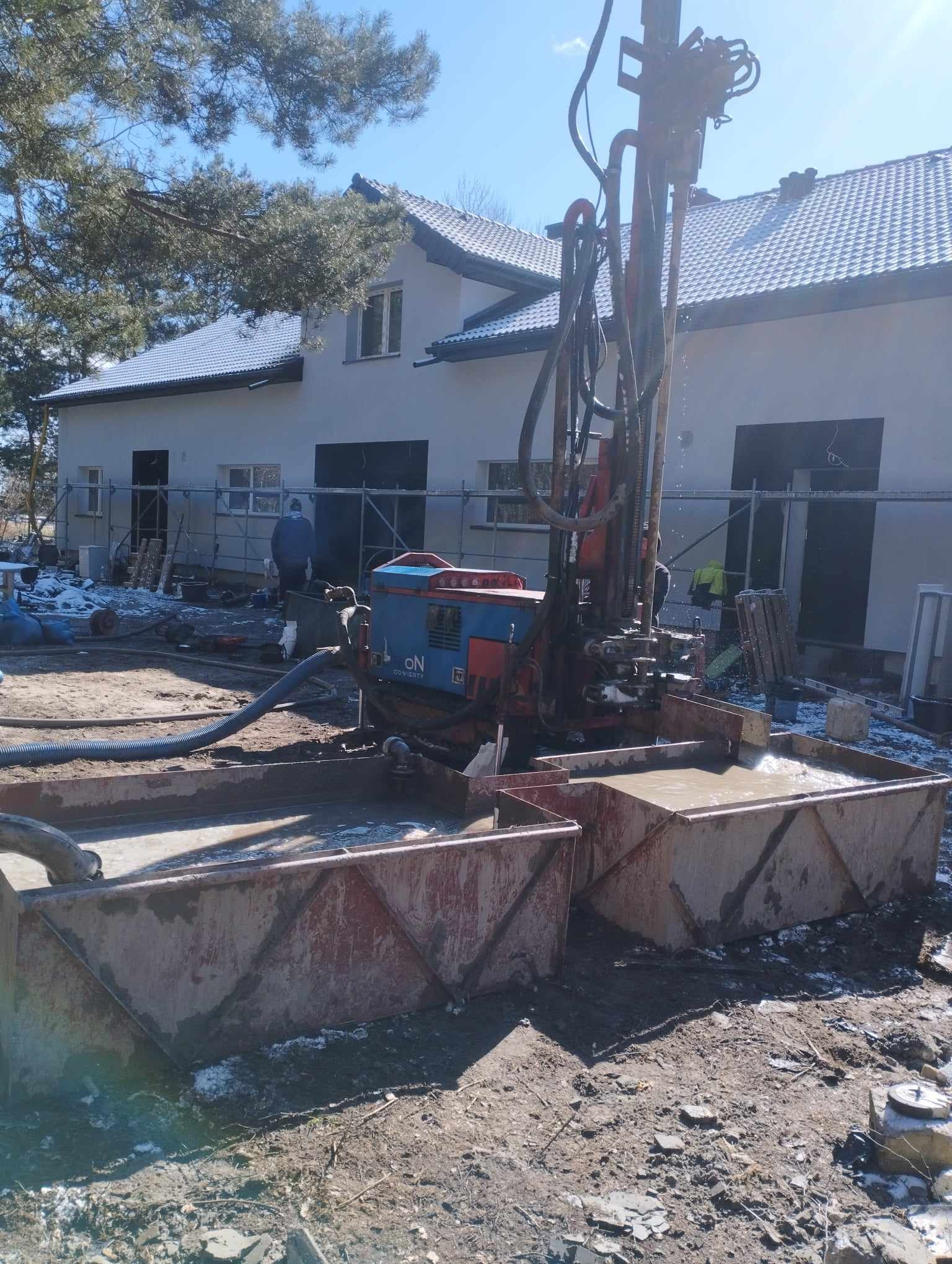 Wiertnica w trakcie pracy, woda wypływa do metalowych zbiorników, w tle budynek w trakcie budowy z rusztowaniem, częściowo ośnieżony teren.