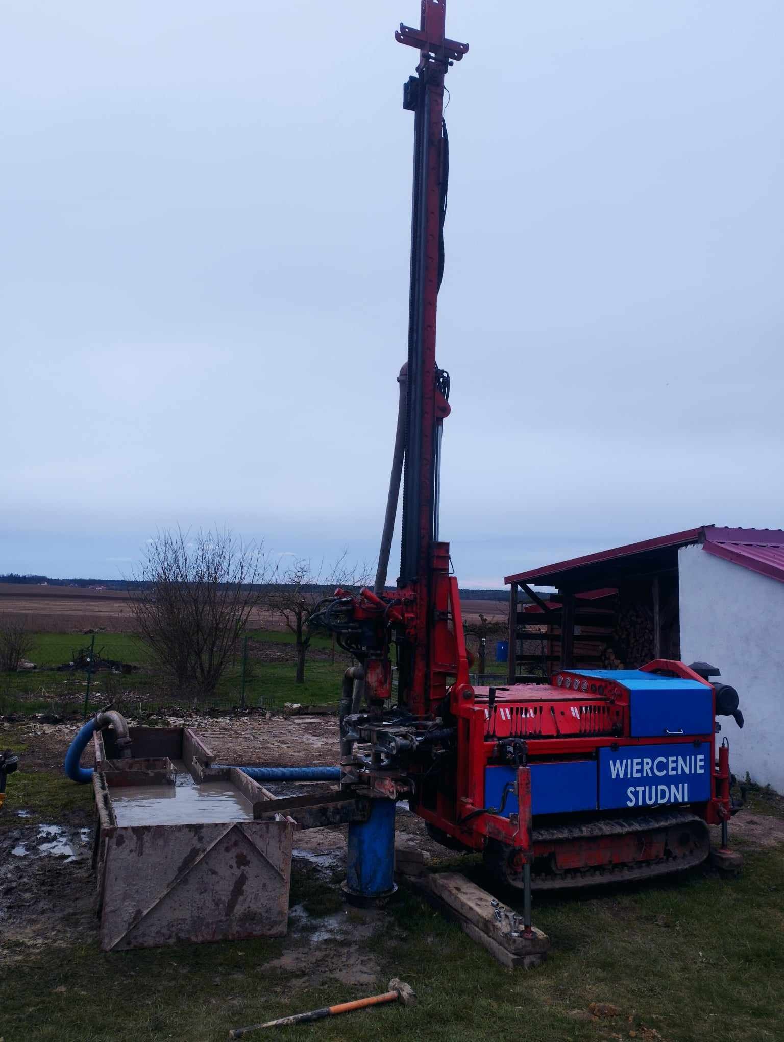 Czerwono-niebieska wiertnica na gąsienicach z napisem 'Wiercenie Studni' na tle pochmurnego nieba i krajobrazu polnego; widoczny zbiornik na wodę i narzędzie na ziemi.