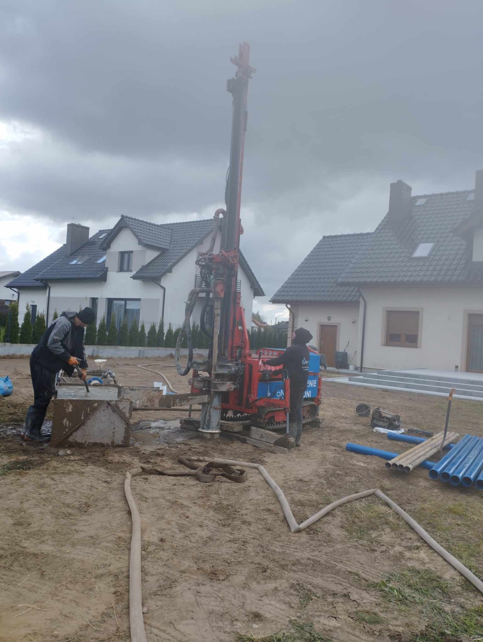 Dwóch pracowników obsługuje czerwoną wiertnicę na podwórku z dwoma domami w tle, widoczne niebieskie rury i węże na ziemi, pochmurne niebo.