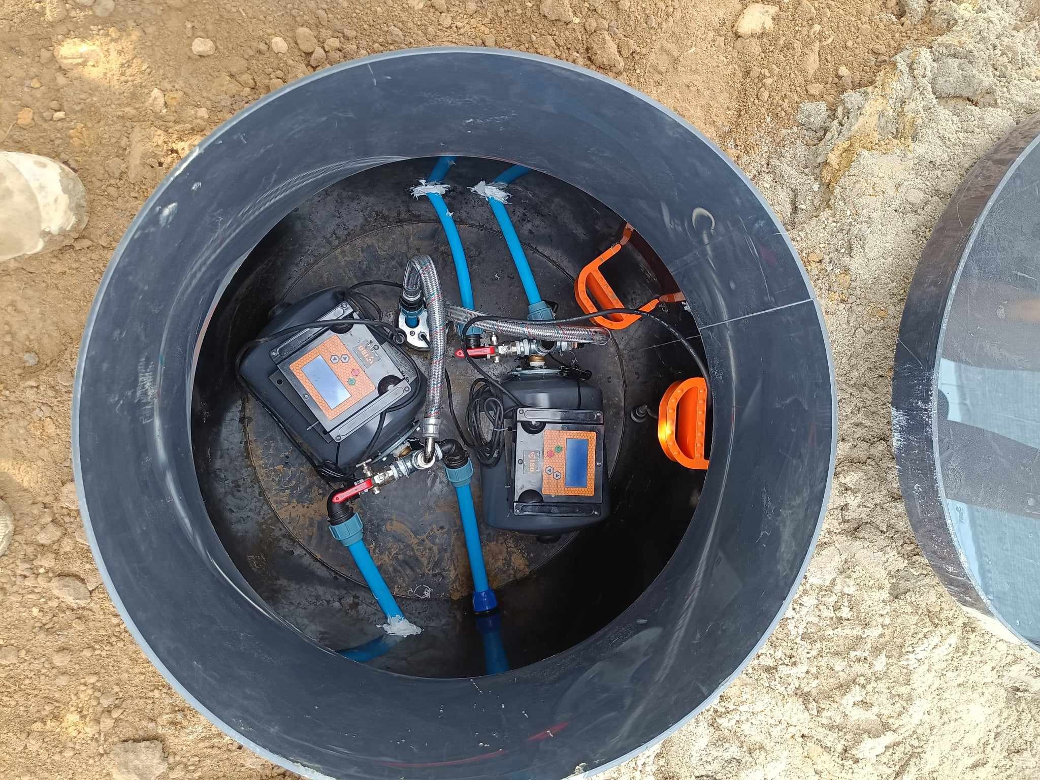Wnętrze studni głębinowej z dwoma pompami i sterownikami, widoczne niebieskie rury i elementy instalacji, otoczone czarnym tworzywem sztucznym, z piaskiem na zewnątrz.