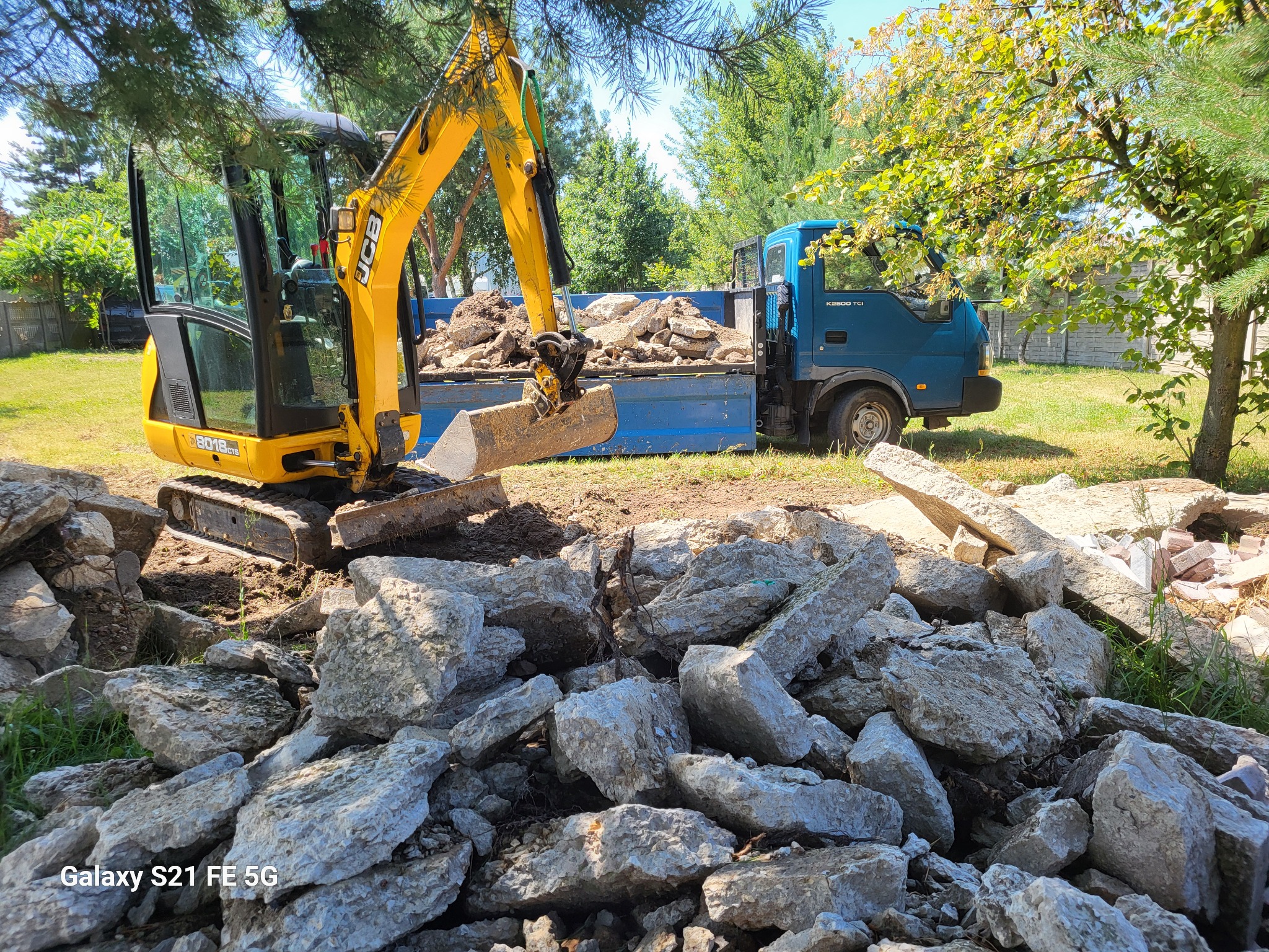 Żółta mini-koparka JCB ładuje gruz betonowy do niebieskiej ciężarówki na tle zieleni drzew i trawy.