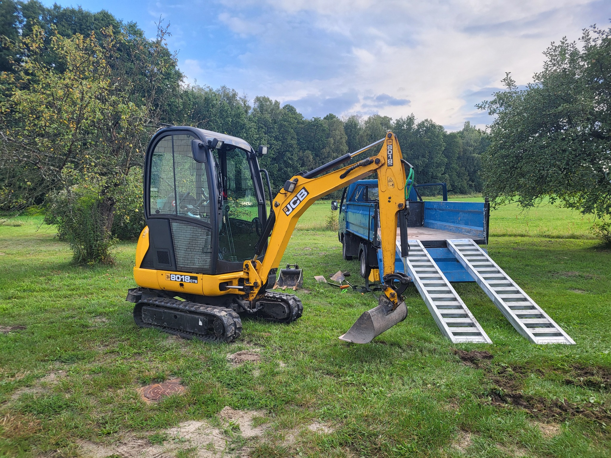Żółta minikoparka JCB 8018 CTS stoi na trawie obok niebieskiej ciężarówki z rampami, przygotowana do transportu lub pracy na działce.