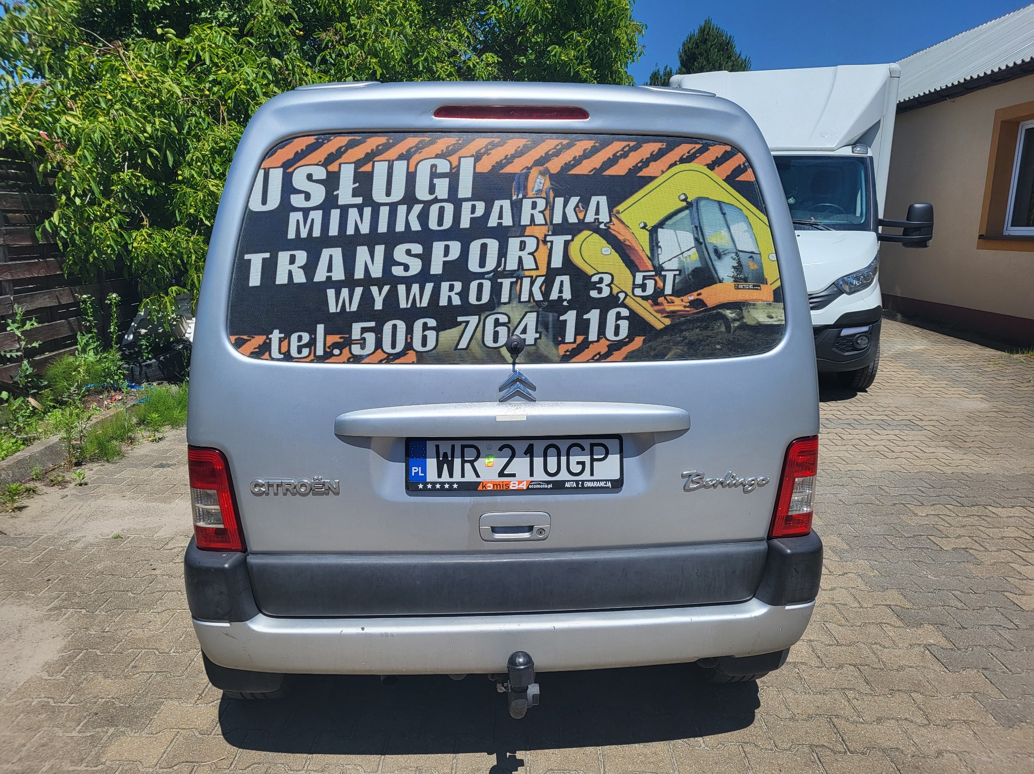 Tył srebrnego Citroena Berlingo z reklamą usług minikoparką, transportu i wywrotki 3,5t, z numerem telefonu na tylnej szybie, zaparkowanego na brukowanym podjeździe, z widocznym białym samochodem...