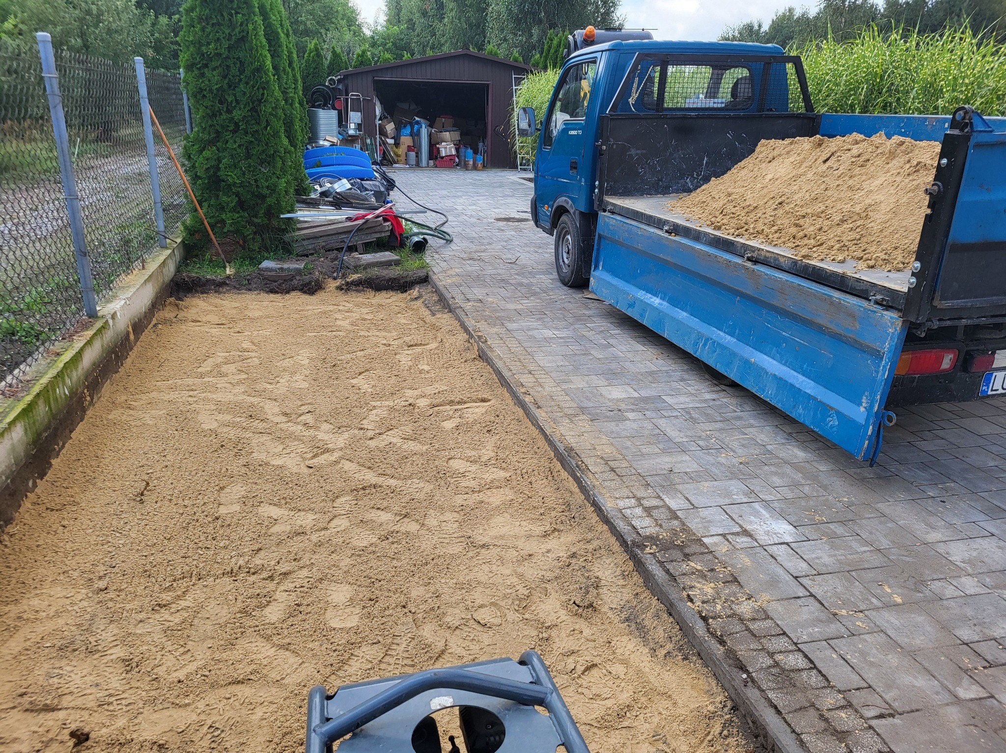 Przygotowanie terenu pod trawnik: wyrównany piasek, ubijarka w tle, niebieska ciężarówka z piaskiem obok kostki brukowej.