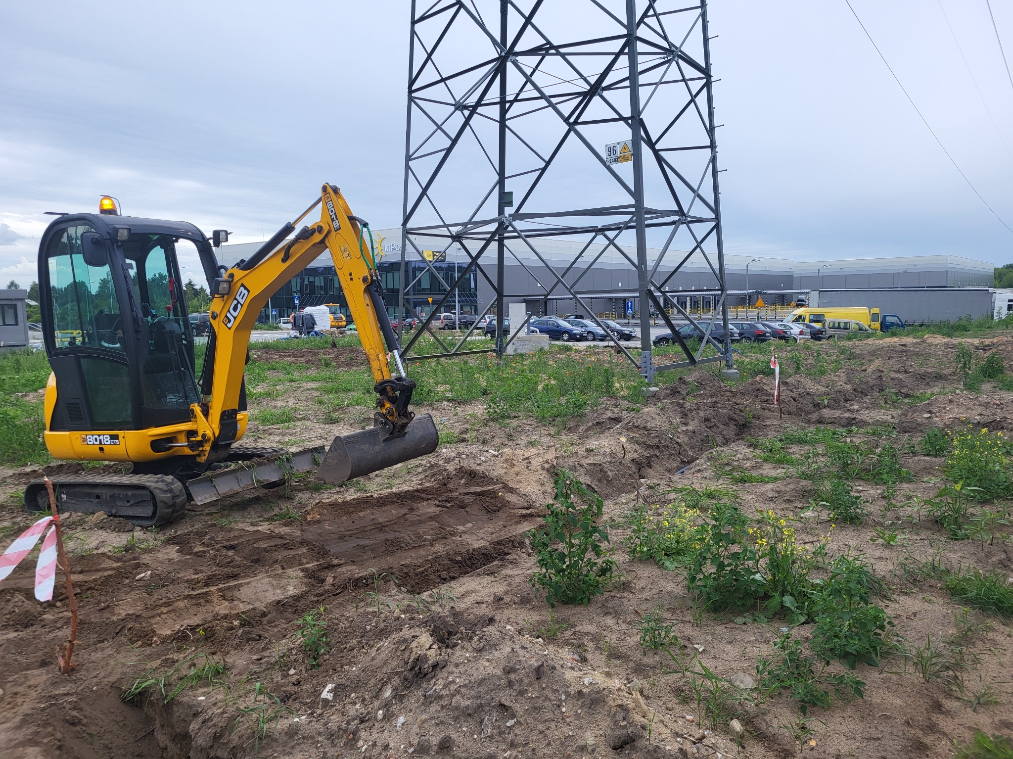 Żółta mini koparka JCB 8018 CTS podczas wykonywania wykopu liniowego w ziemi, z widoczną linią wysokiego napięcia w tle.