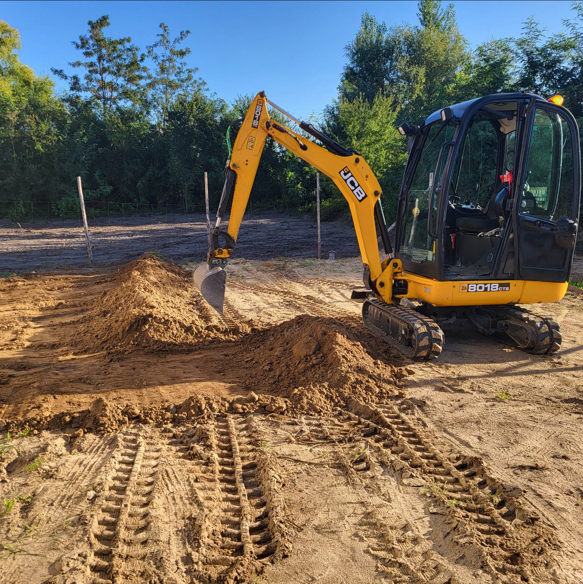 Żółta minikoparka JCB 8018 CTS na gąsienicach, w trakcie prac ziemnych, formuje niewielki nasyp z piasku na placu budowy, widoczne ślady opon, w tle drzewa i częściowo ogrodzenie.
