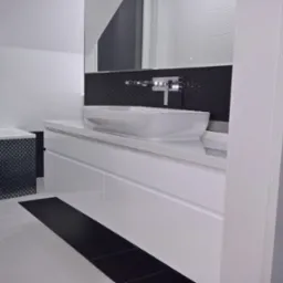 Nowoczesna łazienka z białą, podwieszaną szafką umywalkową z szufladami, owalną umywalką nablatową i baterią ścienną, czarną mozaiką na ścianie oraz fragmentem wanny obłożonej czarno-białą mozaiką.