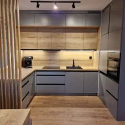 Nowoczesna kuchnia z szarymi szafkami i drewnianymi blatami, z widocznym ekspresem do kawy, czarną armaturą i drewnianą ścianką działową z ceglanym fragmentem ściany w tle.