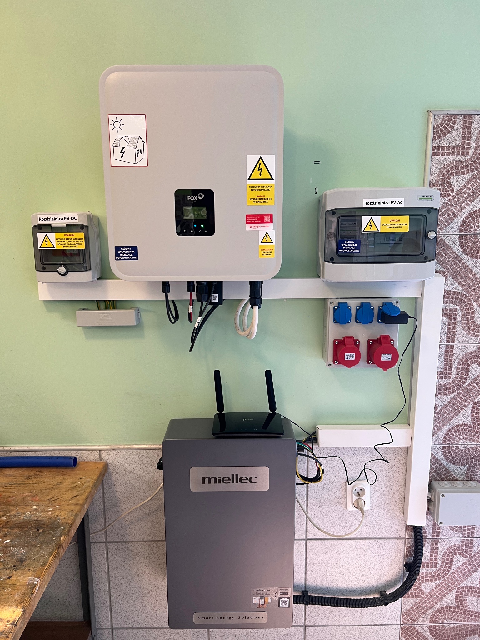 Szara instalacja fotowoltaiczna z rozdzielnicami PV-DC i PV-AC, inwerterem Fox ESS, oraz magazynem energii Miellec, zamontowana na ścianie z płytkami.