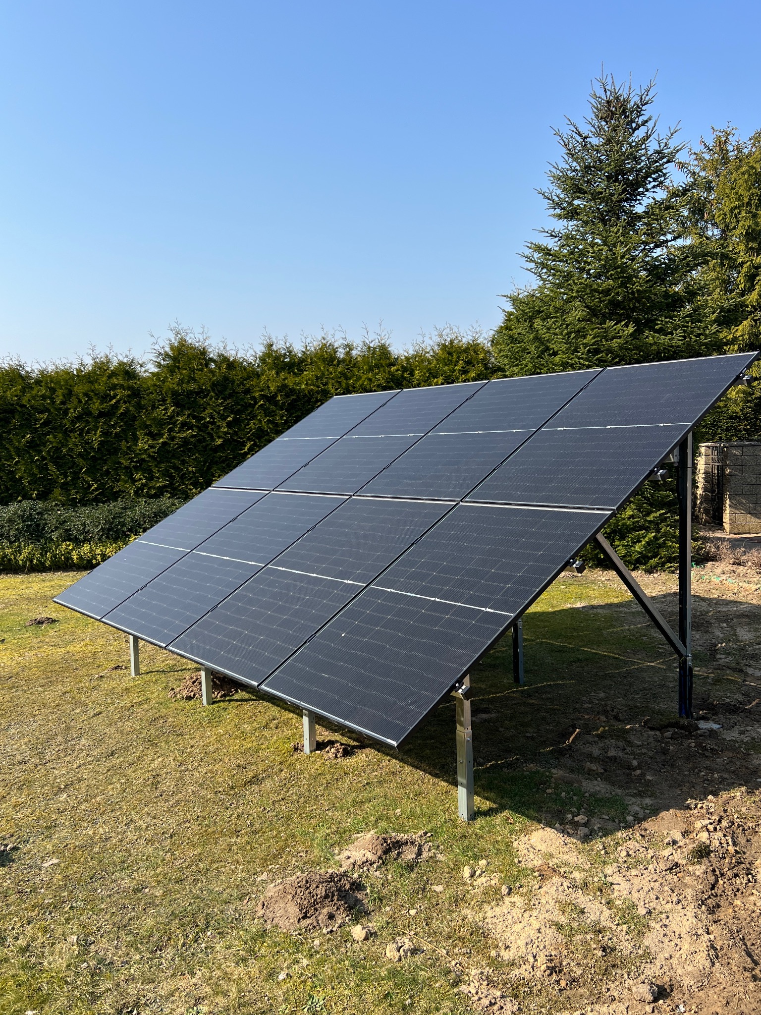 Panel fotowoltaiczny na trawniku, z widocznym montażem i zielonym tłem z drzew i krzewów, na tle błękitnego nieba.
