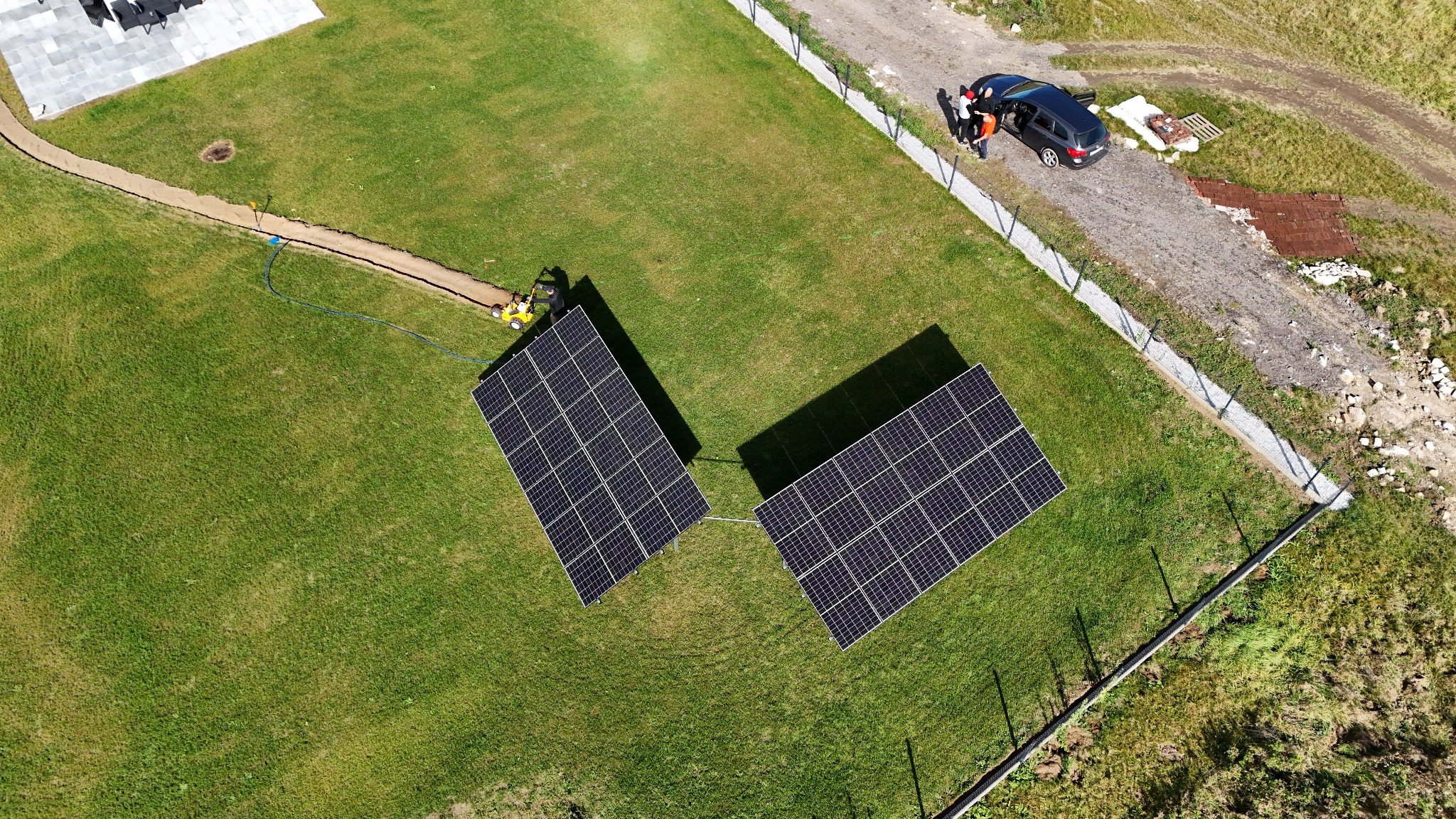 Dwa panele fotowoltaiczne na trawniku, widziane z góry. Obok wykopany rów, a w tle samochód i osoby. Montaż instalacji w toku.