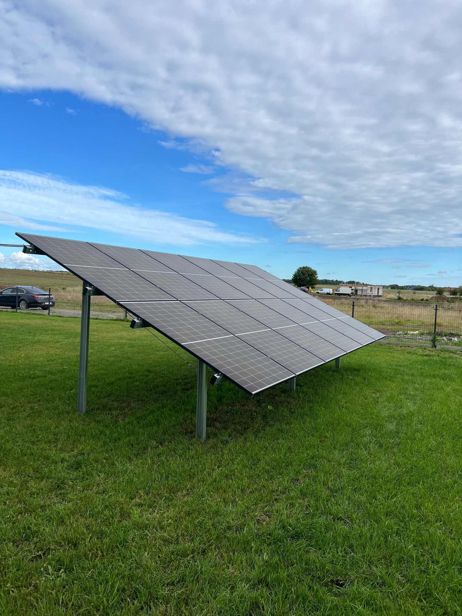 Panel fotowoltaiczny na trawie z widocznym niebem i zabudowaniami w tle, instalacja naziemna, widok z dołu.