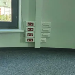 Rząd czterech czerwonych, kwadratowych przycisków alarmowych przeciwpożarowych zamontowanych na białej ścianie obok okna, z szarym dywanem na podłodze.