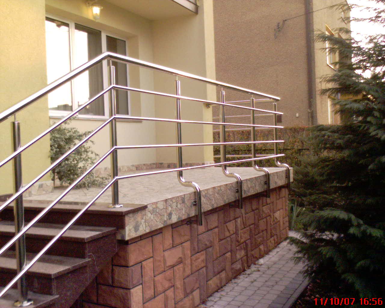 Zewnętrzne schody z brązowego kamienia prowadzące do wejścia z balustradą ze stali nierdzewnej, widok na budynek w tle i krzew po prawej stronie.