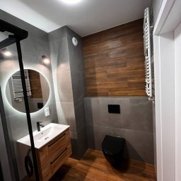 AMIKO GRANDE - Nowoczesna łazienka z drewnopodobną podłogą i płytkami, wisząca toaleta w kolorze czarnym, umywalka z szafką z jasnego drewna, okrągłe lustro z podświetleniem LED.