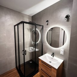 AMIKO GRANDE - Nowoczesna łazienka z kabiną prysznicową w czarnej ramie, okrągłym lustrem z podświetleniem LED i drewnianą szafką pod umywalką, wykończona szarymi płytkami imitującymi kamień.