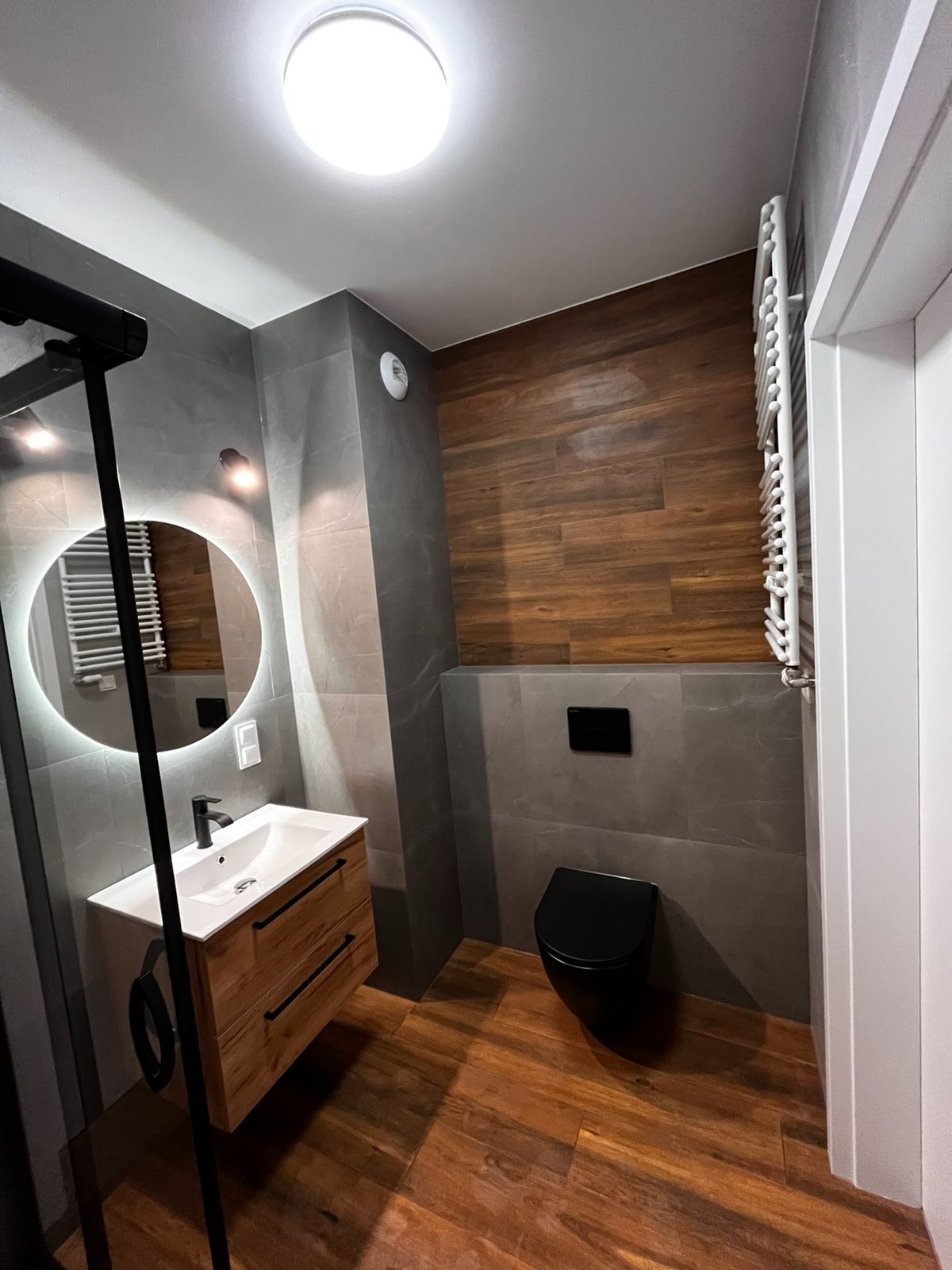 Nowoczesna łazienka z drewnopodobną podłogą i płytkami, wisząca toaleta w kolorze czarnym, umywalka z szafką z jasnego drewna, okrągłe lustro z podświetleniem LED.