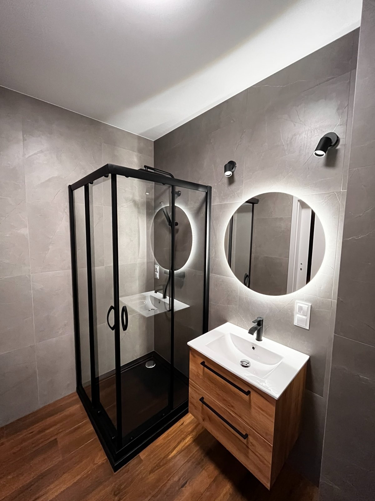Nowoczesna łazienka z kabiną prysznicową w czarnej ramie, okrągłym lustrem z podświetleniem LED i drewnianą szafką pod umywalką, wykończona szarymi płytkami imitującymi kamień.