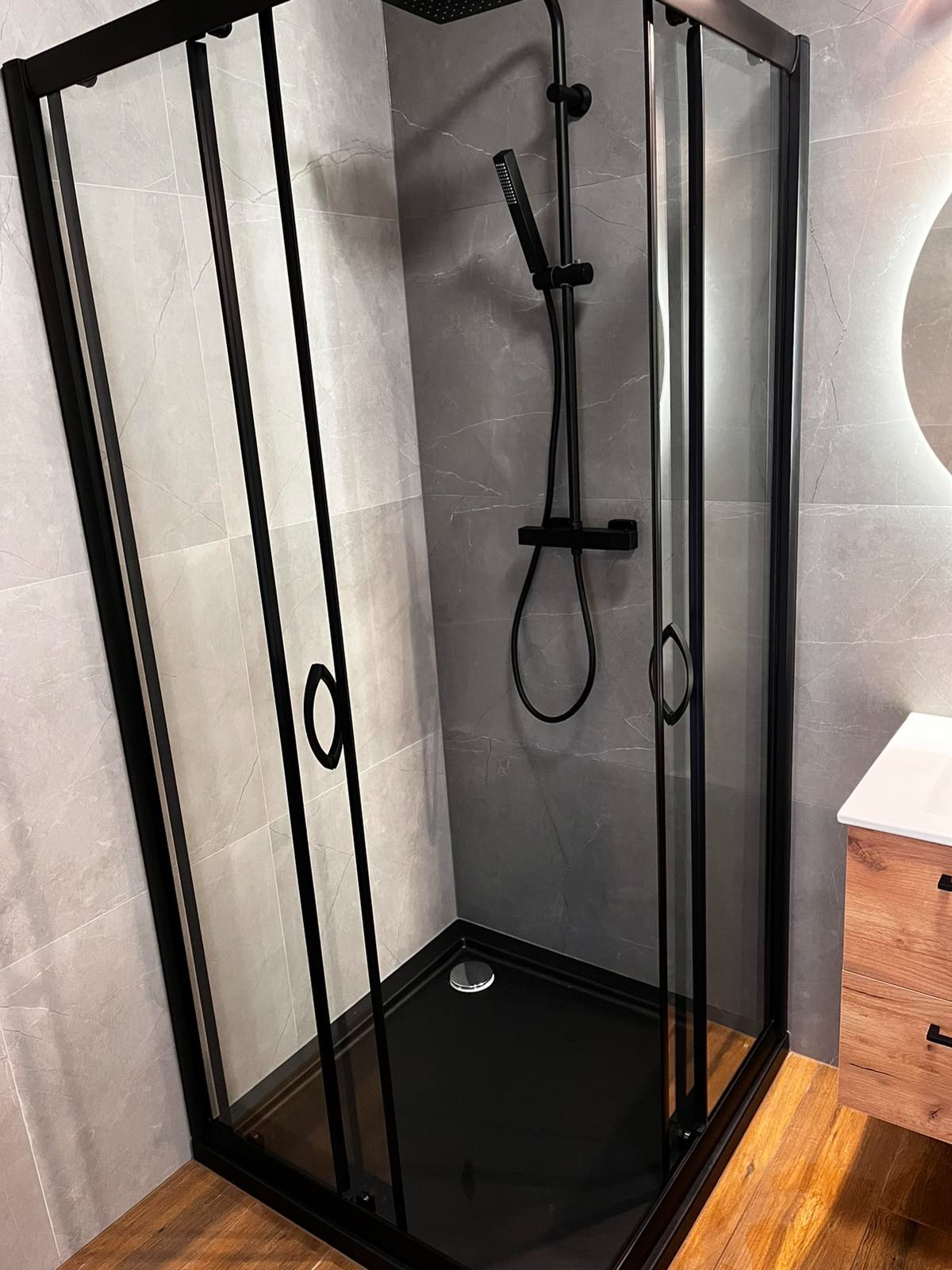 Nowoczesna kabina prysznicowa z czarnymi profilami i deszczownicą, szare płytki imitujące marmur na ścianach, drewnopodobna podłoga, widoczna umywalka z szafką.