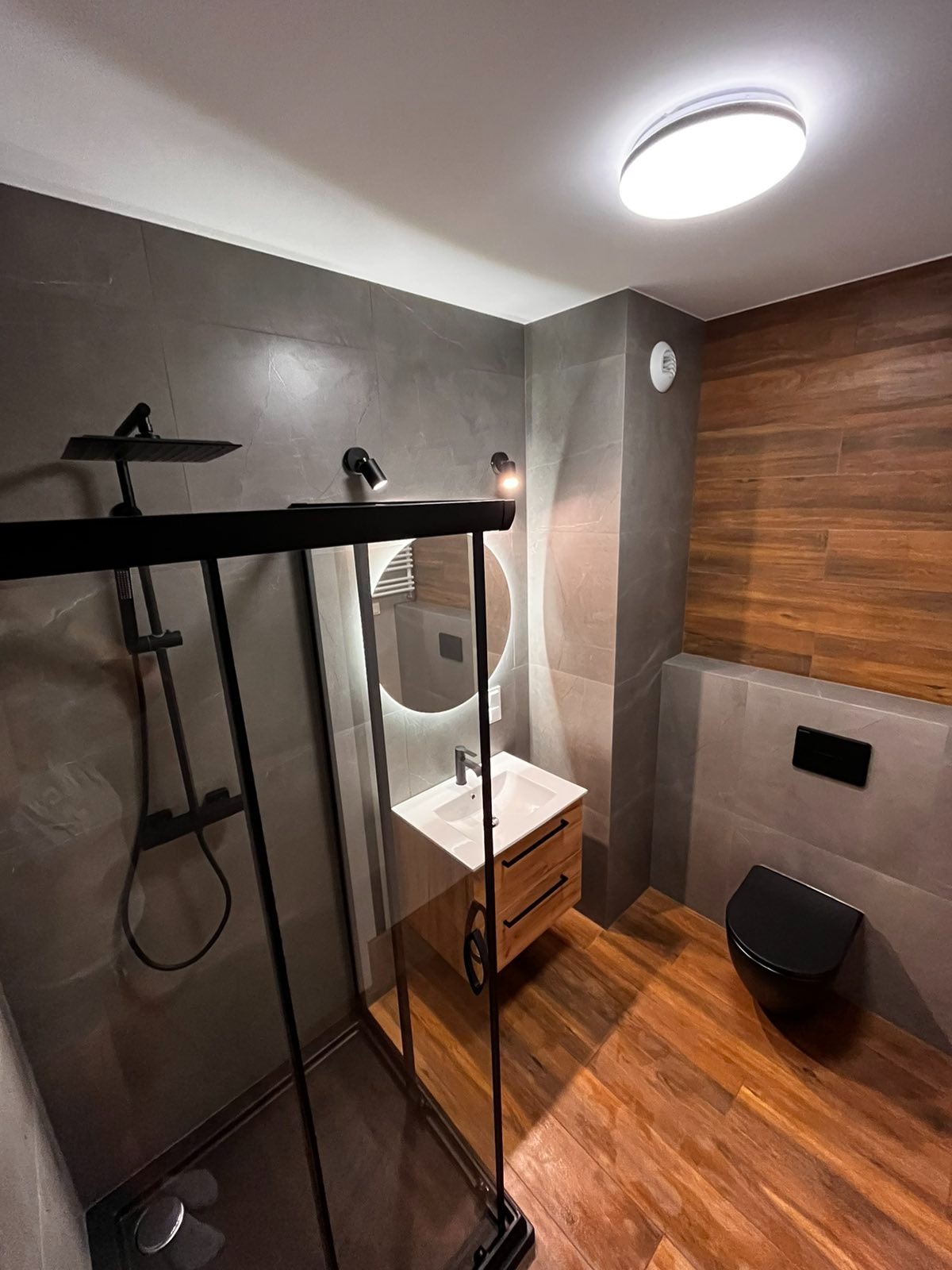 Nowoczesna łazienka z kabiną prysznicową typu walk-in z czarnymi profilami, umywalką z drewnianą szafką, okrągłym lustrem z podświetleniem LED, toaletą w czarnym kolorze i płytkami imitującymi...