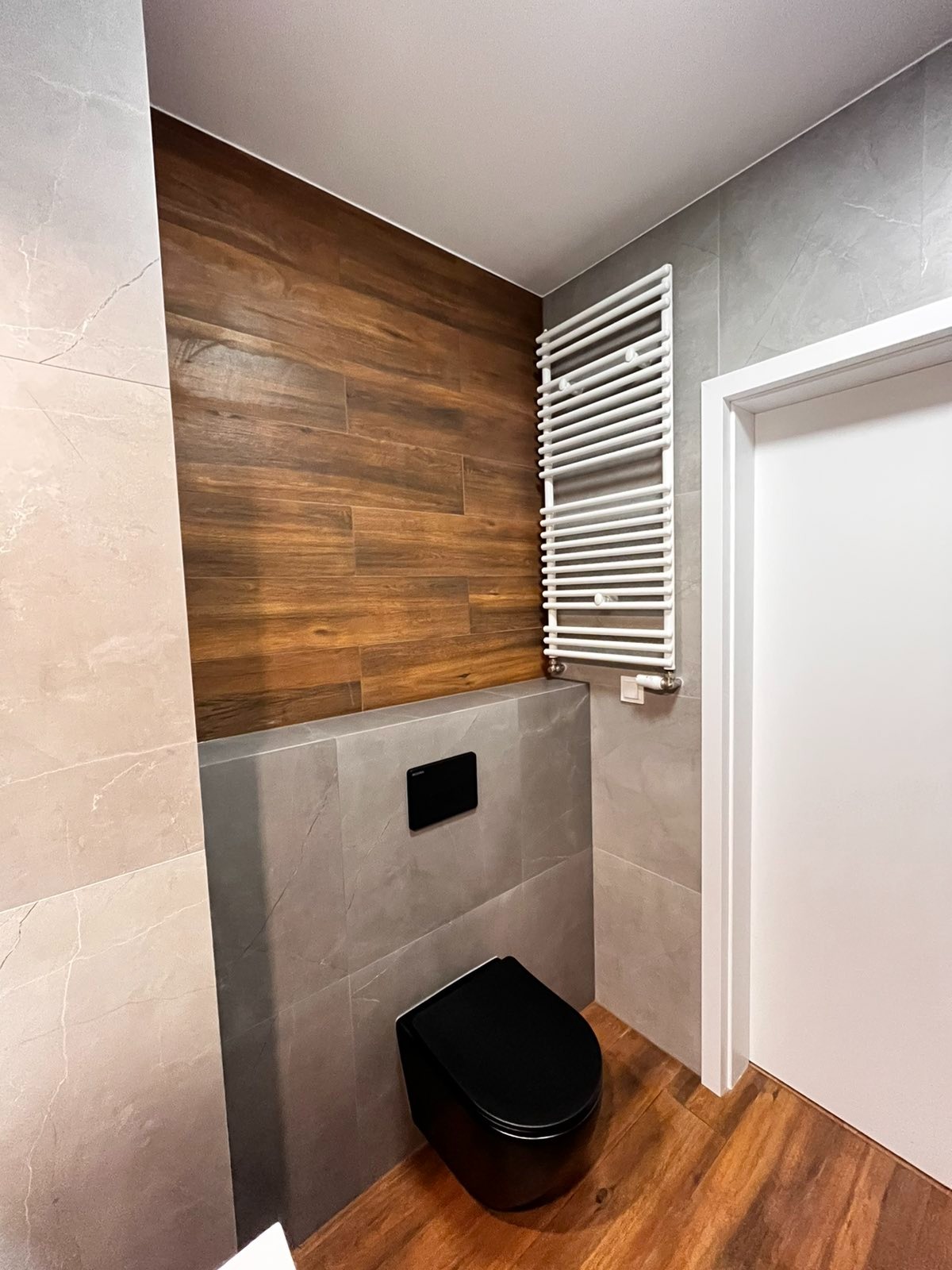 Nowoczesna łazienka z drewnopodobnymi płytkami na ścianie, szarymi płytkami imitującymi kamień, czarną podwieszaną miską WC oraz białym grzejnikiem drabinkowym.