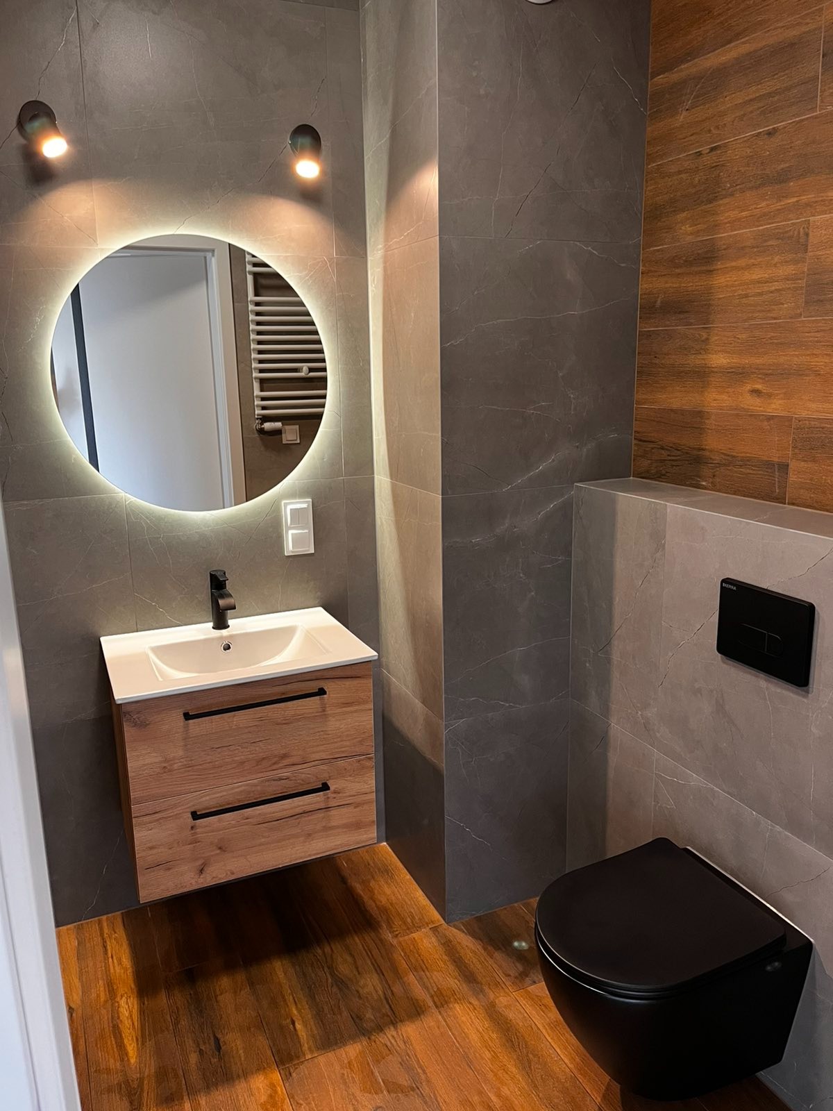 Nowoczesna łazienka z szarymi płytkami imitującymi marmur na ścianach, drewnopodobną podłogą, okrągłym lustrem z podświetleniem LED, umywalką z szafką z drewnianymi frontami i czarną armaturą...