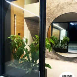 Nowoczesne wnętrze z roślinami w czarnej donicy, widoczne przez przeszklone drzwi, z okrągłym lustrem odbijającym podobny widok, na podłodze logo firmy.