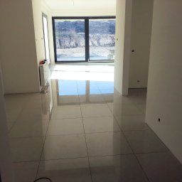 RemontLux - Wnętrze w stanie deweloperskim z jasnymi, błyszczącymi płytkami podłogowymi, świeżo pomalowanymi ścianami na biało i dużym oknem przesuwnym z widokiem na teren budowy.